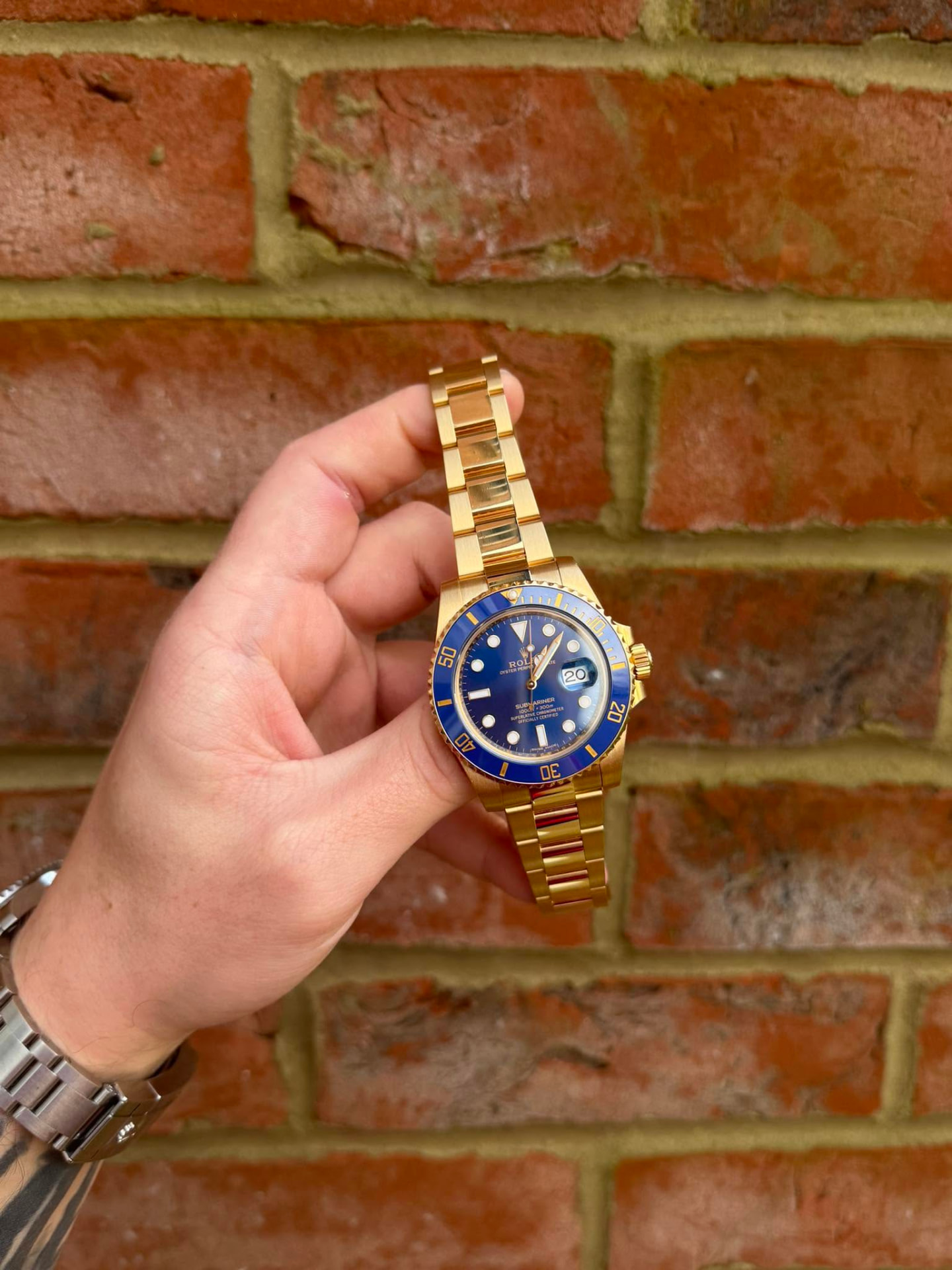 Gold sports watch with a blue dial and bezel held against a brick wall
